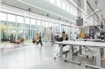 Bildbeschreibung: Eine Gruppe betrachtet Gemälde in einem Ausstellungsraum. Die Gruppe besteht mehrheitlich aus älteren Personen. Eine Frau im Vordergrund stützt sich auf einen Rollator, andere haben sich auf eine Sitzinsel und Klapphocker gesetzt.