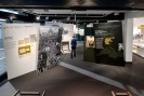 Bildbeschreibung: Der Betrachter blickt von einem erh&ouml;hten Standpunkt in einen Raum mit Ausstellungseinheiten. Auf der farbigen Fotografie sind hintereinander schiefe Stelen angeordnet auf denen viele Texte und Fotografien abgedruckt sind. Als Exponat ist in der Bildmitte ein kleines h&ouml;lzernes Pult zu sehen. In einer Vitrine auf der rechten Seite steht die obere H&auml;lfte einer Schaufensterpuppe, die eine beige Nazi-Uniform tr&auml;gt. Eine Frau geht mit dem R&uuml;cken zum Betrachter in die Raumflucht hinein. Sie wirkt durch die Bewegung auf dem Foto leicht verschwommen.