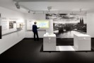 Bildbeschreibung: Die Farbfotografie zeigt die linke Ecke eines Ausstellungsraums. Auf der linken Wand sind in Kopfh&ouml;he Texte und Fotografien &uuml;ber Konrad Adenauer zu sehen. An der rechten Wand ist ein Touchscreen-Bildschirm angebracht, der von einer grauhaarigen, schwarzgekleideten Frau ausprobiert wird. Daneben h&auml;ngt eine gro&szlig;e Schwarzwei&szlig;-Fotografie. Die Fotografie zeigt eine historische Aufnahme der K&ouml;lner Skyline mit Blick auf den K&ouml;lner Dom und die Hohenzollernbr&uuml;cke. In der Mitte des Raumes stehen zwei h&uuml;fthohe wei&szlig;e Vitrinen.