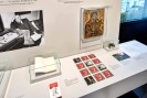 Bildbeschreibung: Die Farbfotografie l&auml;sst den Betrachter auf eine wei&szlig;e Ausstellungseinheit blicken. Auf dem Tisch liegt links ein aufgeschlagenes Buch in einem Glaskasten, darunter liegt ein geschlossens Buch, durch das der Besucher bl&auml;ttern kann. In der Mitte liegen in vier Reihen je drei quadratische farbige Bilder nebeneinander. Es ist ein Memoryspiel und auf den rotgrundigen Deckbl&auml;ttern ist eine Zeichnung von Conny dem Fuchs zu sehen. Rechts daneben steht fast an der Wand eine kleine Glasvitrine mit einem Spielzeugtier darin. Dar&uuml;ber, an der Wand h&auml;ngt eine Ikone der Mutter Gottes, die auf ihren gekreuzigten Sohn Jesus blickt. Links an der Wand h&auml;ngt eine Schwarzwei&szlig;-Fotografie auf der der sitzende Konrad Adenauer zu sehen ist.