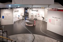 Bildbeschreibung: Auf drei großen Ausstellungswänden, die sich um eine Sitzinsel gruppieren steht: „Was verbindet uns?“, „Was bewegt uns?“ und „Was lieben wir?“. An den Ausstellungswänden sind Texte und Vitrinen mit Objekten angebracht.